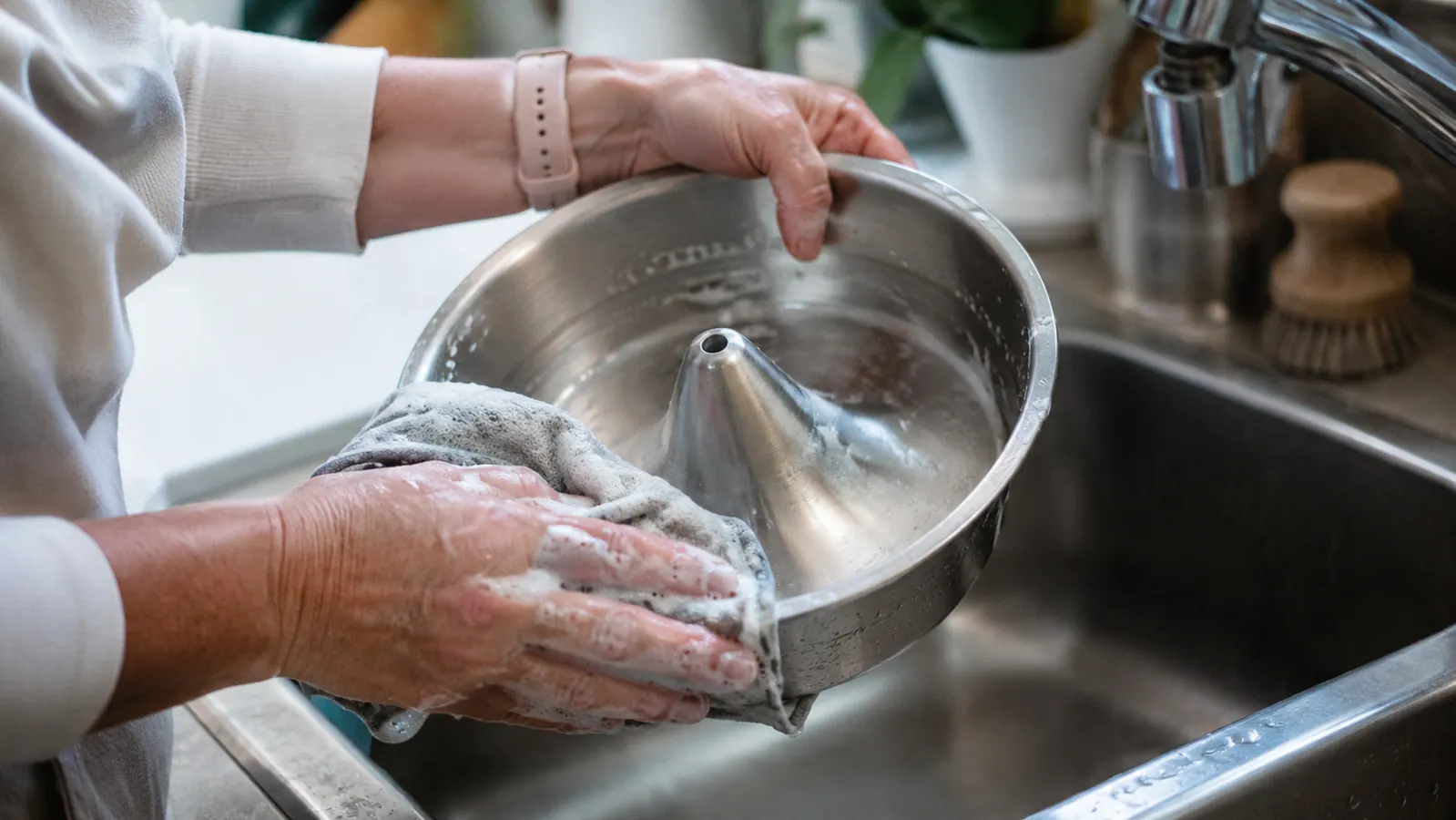 Hands scrubbing the stainless steel bowl of a pet fountain at a kitchen sink with a cloth
